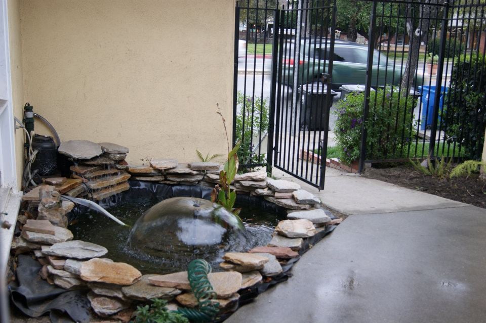 The entrance courtyard, a stone-ringed pond with a dome fountain beside a yellow wall, with an iron gate open to the street