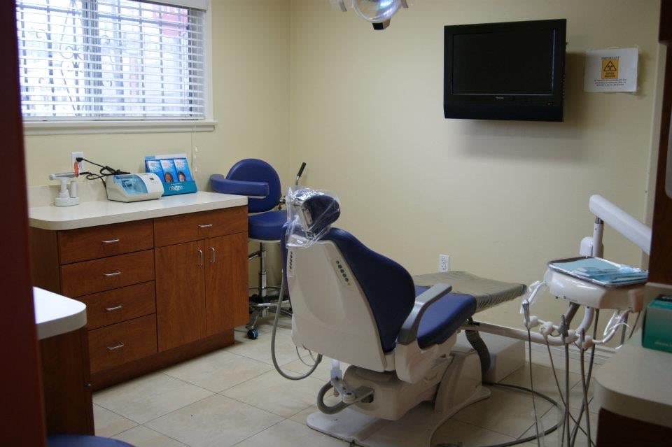 A treatment room with a blue dental chair, wood cabinets and daylight through the blinds